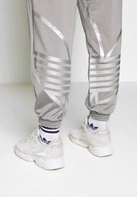 Šedé kalhoty s reflexními stříbrnými geometrickými vzory nad bílými sportovními teniskami a bílými ponožkami Adidas s černým logem a pruhy.