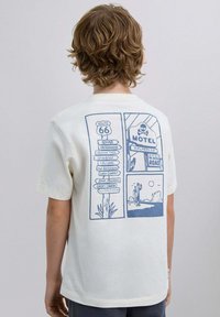 Camiseta blanca de algodón con un diseño gráfico azul en la parte posterior que presenta referencias a la señal de Route 66 y una ilustración de un paisaje desértico.