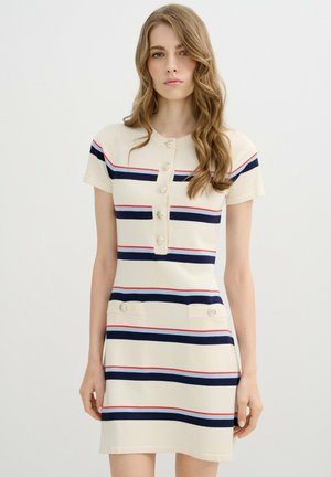 Joven mujer con cabello largo y ondulado que lleva un vestido corto de manga corta color crema con rayas horizontales azul marino y rojas, botones delanteros y solapas de bolsillos abotonadas.