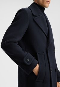 Marineblauer Wollmantel mit maßgeschneidertem Design, doppelt geknöpftem Vorderteil, großem Reverskragen und geknöpften Manschetten. Verfügt über seitliche Taschen und eine weiche Textur.