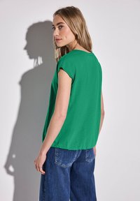 Zelený top s krátkým rukávem, volného střihu a kulatým výstřihem, v kombinaci s modrými širokými džíny. Jednoduchá textura, minimální detaily.
