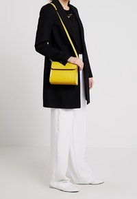 Borsa a tracolla gialla in pelle con un motivo geometrico e chiusura a patta, indossata con un cappotto nero e pantaloni bianchi a gamba larga.