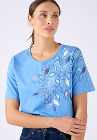 Mujer con una camisa de manga corta azul claro con un patrón abstracto de hojas colorido, mirando hacia la derecha, sosteniendo su brazo izquierdo con su mano derecha.