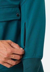 Hand die de mouwzak van een blauwe jas met een geribbelde manchet en een klepzak met een zwarte knoop op het bovenste deel van de arm dichtritst.