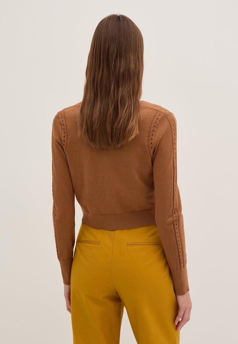 Braun gestrickter Pullover mit einem verkürzten Design und geripptem Saum, der dekorative Perforationen an den Ärmeln aufweist. Kombiniert mit gelben Hosen.