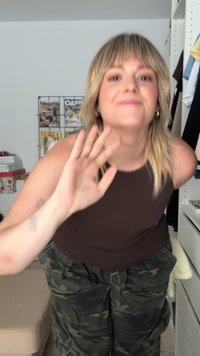 Bruine mouwloze top, camo cargobroek, golfgebaar. Achtergrond omvat een plank met tijdschriften en kleding. Neutrale binnenruimte.