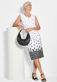 Weißes ärmelloses Kleid mit schwarzen Blattmustern und einem blumigen Saum, kombiniert mit einer schwarzen Halbmondtasche und schwarzen Blockabsatzsandalen.