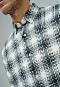 Camisa de cuadros en blanco y negro, diseño de botonadura con cuello, que presenta una tela texturizada y patrones horizontales y verticales distintivos.