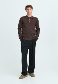 Brauner Strick-Polo-Pullover mit Kragen und einer Drei-Knopf-Leiste, kombiniert mit lockeren schwarzen Hosen und beigen Schuhen. Glatte Textur im gesamten Design.