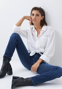 Camisa blanca de botones con mangas arremangadas, jeans ajustados de mezclilla azul y botas de tacón negro hasta el tobillo, posando frente a un fondo uniforme.