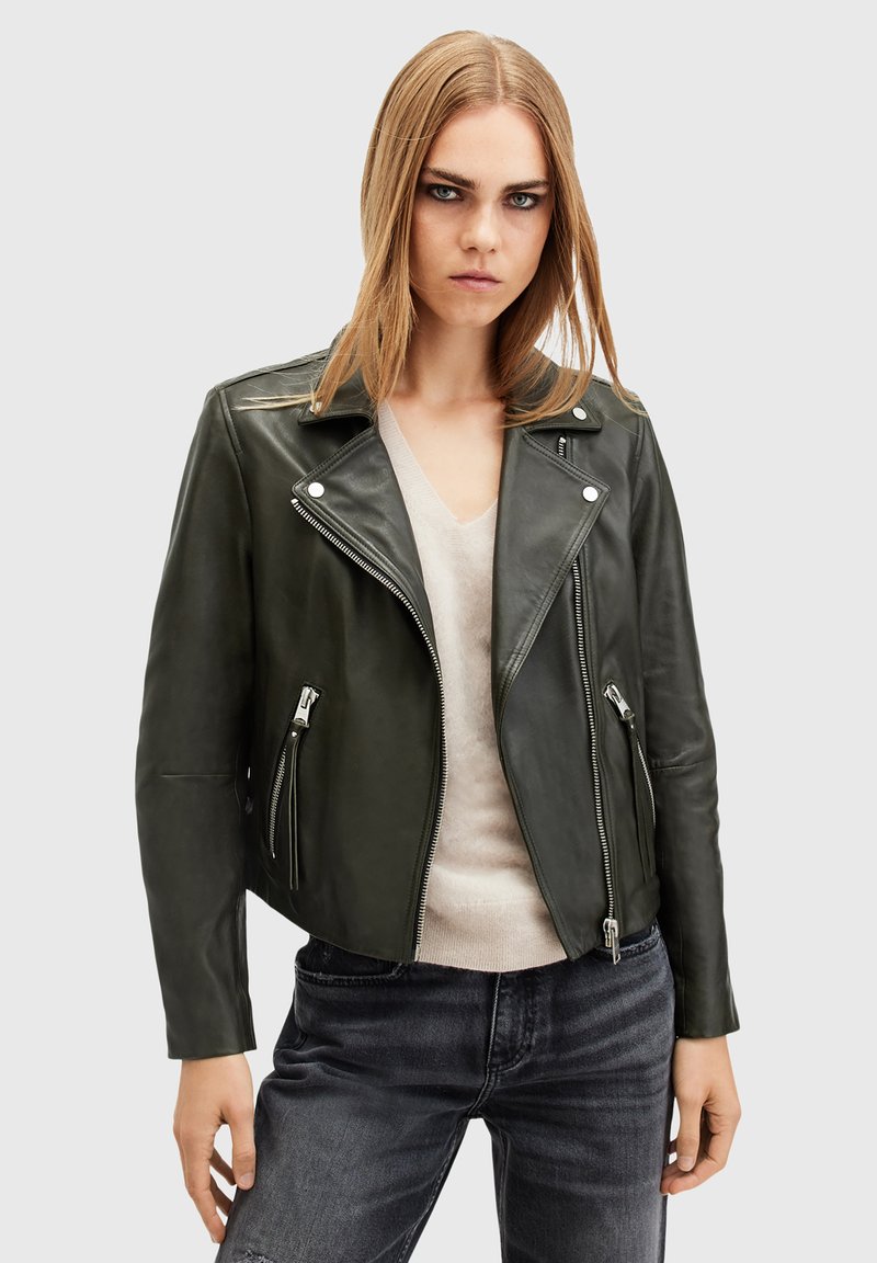 Giacca da moto in pelle verde scuro con hardware argentato, tasche con zip e un ampio revers. Indossata sopra un maglione chiaro e jeans.
