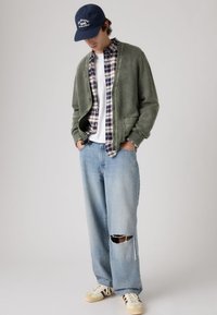 Νεαρός άνδρας με ναυτικό καπέλο, πράσινη ζακέτα, καρό πουκάμισο, σκισμένο ανοιχτό μπλε τζιν και μπεζ αθλητικά παπούτσια, στέκεται με τα χέρια στις τσέπες, το κεφάλι κάτω.