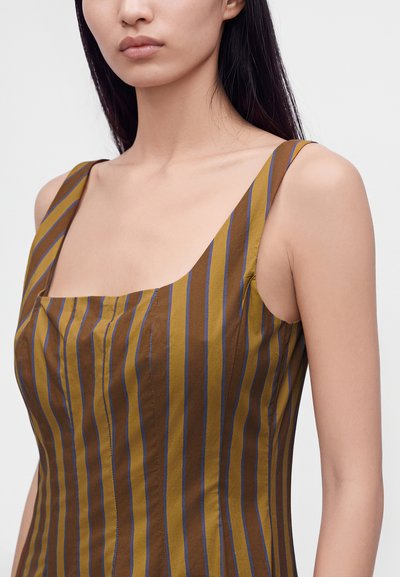 Femme portant une robe marron sans manches avec des rayures verticales bleues et jaunes, montrant le cou, la clavicule et la partie supérieure de la poitrine.