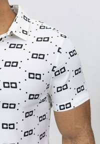 Chemise à manches courtes en tissu blanc avec un motif géométrique noir répétitif et de petits pois noirs. Présente un col classique et des manches à revers.