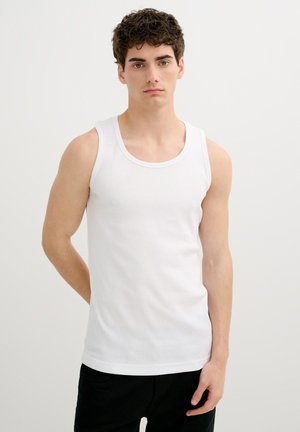 Joven con cabello rizado, vestido con una camiseta sin mangas blanca y pantalones negros, de pie frente a un fondo blanco liso.