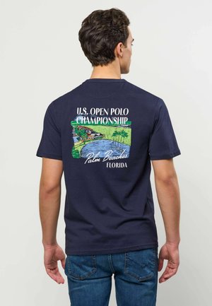 Rövid sötét hajú férfi sötétkék inget visel, melyen színes U.S. Open Polo Championship Palm Beaches Florida grafikája látható a háton.