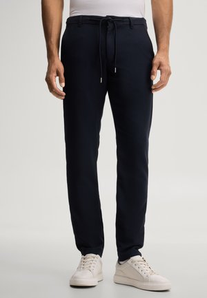 Man die zwarte broek met trekkoord en witte sneakers draagt, staande tegen een effen achtergrond.