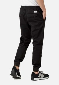 Pantalones jogger negros con cintura elástica, dos bolsillos traseros y puños acanalados, combinados con zapatillas negras y blancas sobre un fondo claro.