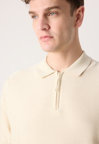 Beige zip-up piquétröja med en texturerad stickad finish. Har en klassisk krage och en enkel framdragkedja, lämplig för avslappnad klädsel.