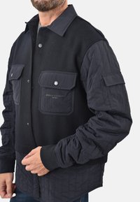 Giacca nera con maniche trapuntate, bottoni a pressione e tasche sul petto doppie. Mix di materiali in lana e nylon, con una sezione inferiore testurizzata.