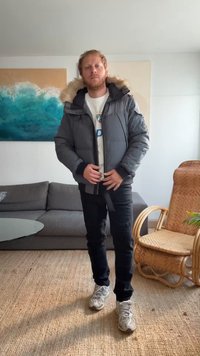 Casaco puffer cinza com capuz de pelo sintético, fecho frontal com zíper e detalhes em preto nos punhos. Conjunto casual combinado com jeans pretos e ténis cinzentos.
