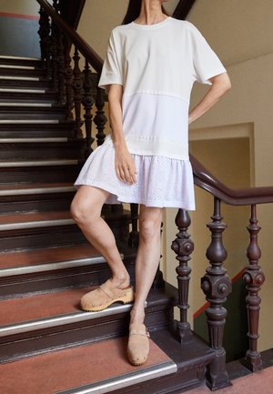 Femme debout sur un escalier en bois portant un haut blanc à manches courtes avec une jupe ajourée et des sabots en daim beige ornés de clous.