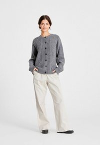 Femeie stând cu mâinile în buzunare, îmbrăcată cu o cardigan tricotat gri cu nasturi, pantaloni bej largi și pantofi negri fără toc, pe un fundal simplu.