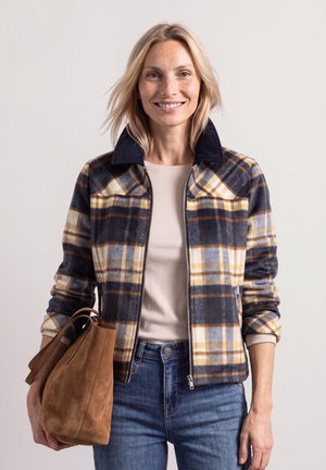 Smilende kvinde med blondt hår iført en ternet jakke, beige top, blå jeans og med en brun ruskindshåndtaske ved siden af sig.