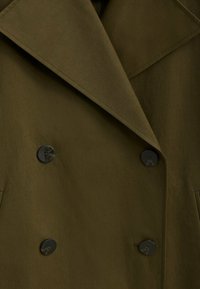 Manteau croisé vert olive avec de grands revers et deux boutons noirs. Tissu lisse et texturé avec des poches latérales.