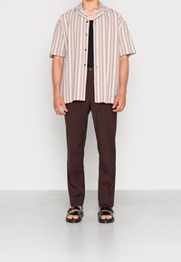 Korte mouwen, een knoopjesoverhemd met horizontale crèmewitte en mauve strepen, gecombineerd met donkerbruine broeken en zwarte sandalen.