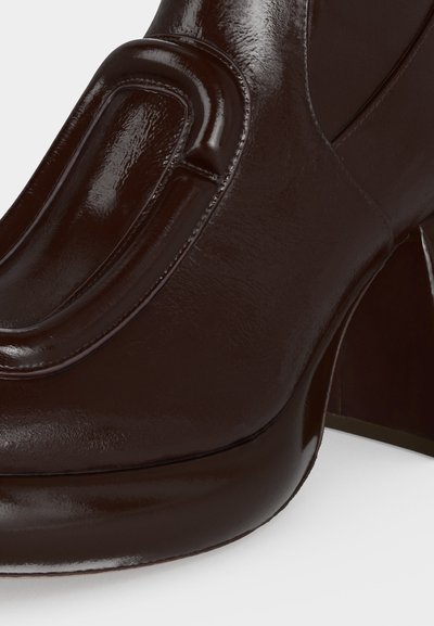 Gros plan d'une chaussure à talon haut en cuir brun foncé brillant, avec un bout rond et des détails de couture décorative.