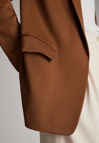 Brązowa marynarka o dopasowanym kroju, z pojedynczą kieszenią z przodu i gładką fakturą materiału. Projekt ma nowoczesny wygląd.