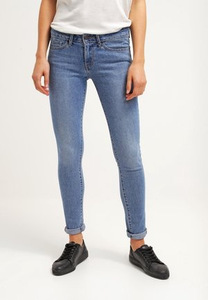 Femme portant un jean skinny bleu clair retroussé, des baskets noires et une chemise blanche, debout devant un fond uni.