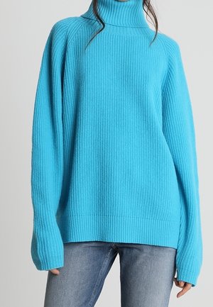 Pull col roulé turquoise avec texture côtelée, coupe décontractée et manches longues, associé à un jean bleu.
