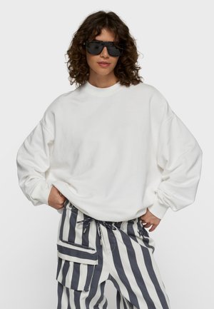 Kvinde med krøllet hår iført oversize hvid sweatshirt, sorte solbriller og sort-hvide vertikale stribede bukser med cargolommer.