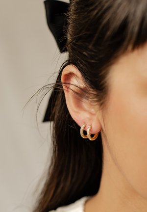Goldene Creolen mit gebogenem Design, die eine Reihe von kleinen klaren Steinen als Akzent tragen. Die Haare sind mit einer schwarzen Bänder-Schleife gestylt.