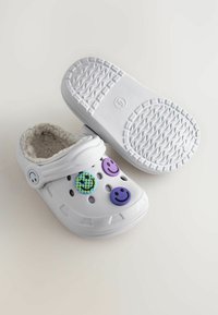 Weiße Clogs mit flauschigem Innenfutter, ausgestattet mit drei dekorativen Knöpfen: zwei mit lächelnden Gesichtern und einem gemusterten Kreis. Strukturiert, rutschfeste Sohle.