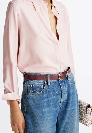 Kobieta ubrana w jasnoróżową bluzkę zapinaną na guziki, włożoną do niebieskich dżinsów z brązowym skórzanym paskiem, trzymająca beżową torebkę w wzory.