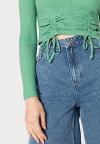 Grünes, geripptes Langarmoberteil mit Raffung und Bändern an der Taille, kombiniert mit hoch taillierten blauen Jeans aus Denim mit Knopfverschluss.