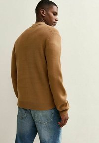 Uomo che indossa un maglione marrone a maniche lunghe e jeans di denim blu, in piedi e che guarda di lato contro uno sfondo neutro.