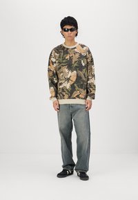 Maglione con stampa mimetica in tonalità di verde, marrone e accenti crema. Vestibilità ampia, polsini a coste, abbinate a jeans azzurri e calzature nere.