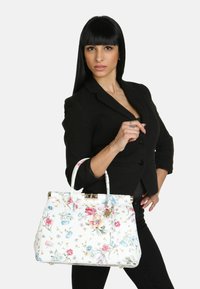 Hvid struktureret håndtaske med blomstermønster i pink, blå og gul. Har en klaplukning og korte håndtag. Model iført sort blazer.