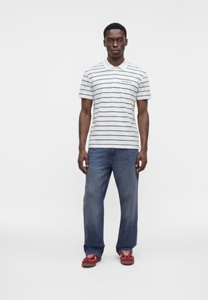 Mann steht frontal, trägt ein weiß gestreiftes Poloshirt, blaue Jeans und rote Turnschuhe vor einem schlichten, hellen Hintergrund.