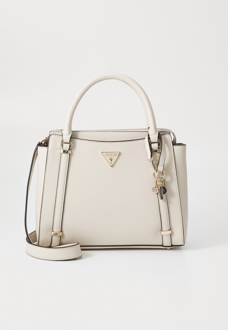 Sac à main Guess couleur crème avec deux poignées, une bandoulière amovible et des détails logo et breloque dorés sur fond blanc.