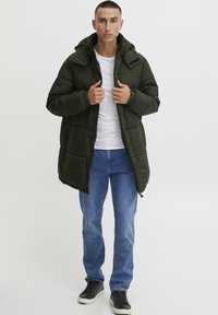 Mørkegrønn boblejakke med hette, kviltet design, frontglidelås og elastiske mansjetter. Brukt over en hvit skjorte og blå jeans.
