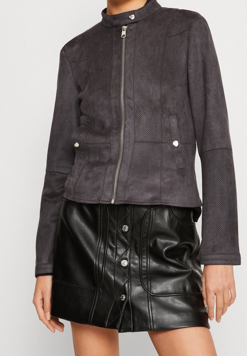 Giacca grigia in finta pelle scamosciata con chiusura frontale con zip, colletto corto e pannelli traforati, abbinata a una mini gonna in pelle nera con dettagli a bottoni.