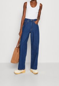 Woman wearing white tank top, high-waisted wide-leg blue jeans, beige clogs, holding a large tan suede tote bag, standing against a white background.