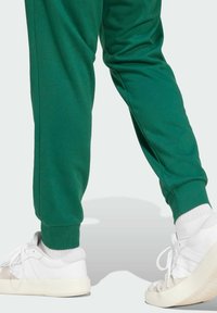 Pantalones deportivos verdes con puños ajustados, combinados con zapatillas blancas que tienen suelas y cordones texturizados. Textura de tela suave visible.