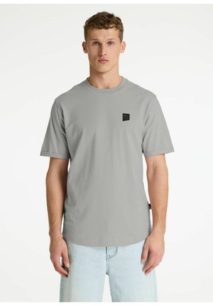 Mladý muž s krátkymi kučeravými vlasmi, oblečený v svetlosivej tričku s krátkymi rukávmi a svetlomodrých džínsoch, stojaci pred jednoduchým bielym pozadím.