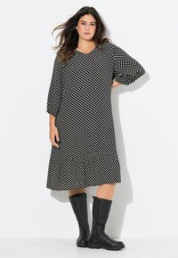 Schwarzes Kleid mit weißen Punkten, mit tiefem Taillenschnitt und ausgestelltem Saum. Hat 3/4-Ärmel. Kombiniert mit knielangen schwarzen Lederstiefeln.